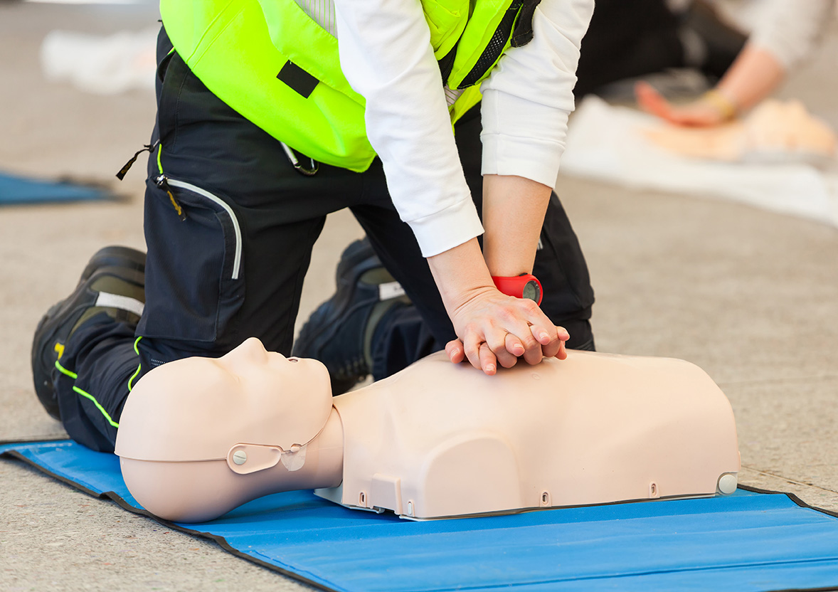 A person wearing a high-visibility vest practices CPR on a training mannequin, demonstrating proper chest compression technique during a first aid training session.