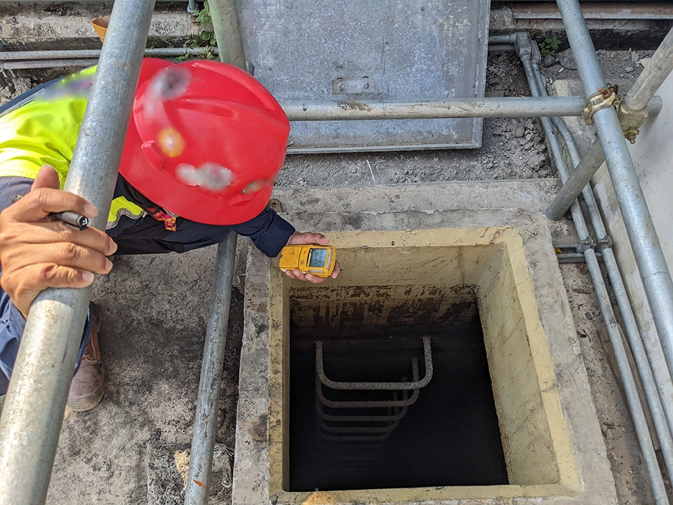 Worker wearing a red helmet and high-visibility vest inspecting a confined space using a gas detection device near a square entry point with a fixed ladder.