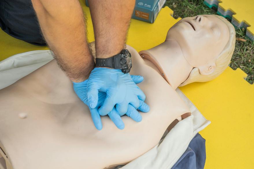 Close-up of hands wearing blue gloves performing CPR chest compressions on a training mannequin. The scene depicts a first aid or CPR training session, emphasizing life-saving techniques and proper procedure.
