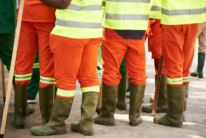 Workers in high-visibility safety uniforms and rubber boots standing together on a construction site. The bright orange and yellow gear highlights safety precautions in an outdoor work environment.