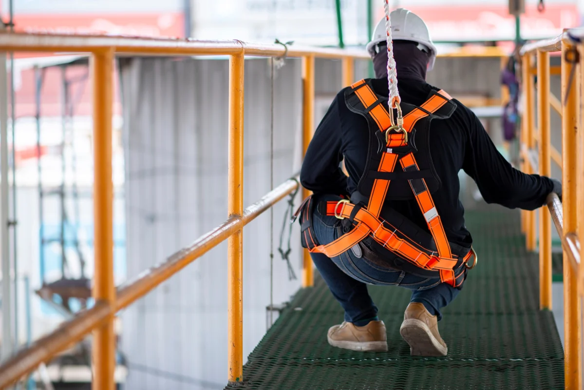 Worker wearing a safety harness and hard hat, crouching on an elevated walkway with safety railings, highlighting fall protection measures in a construction or industrial setting.