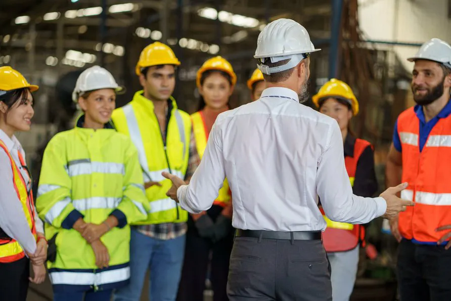 Supervisor in a white hard hat leading a safety briefing with a diverse group of workers wearing high-visibility gear and helmets inside an industrial facility.