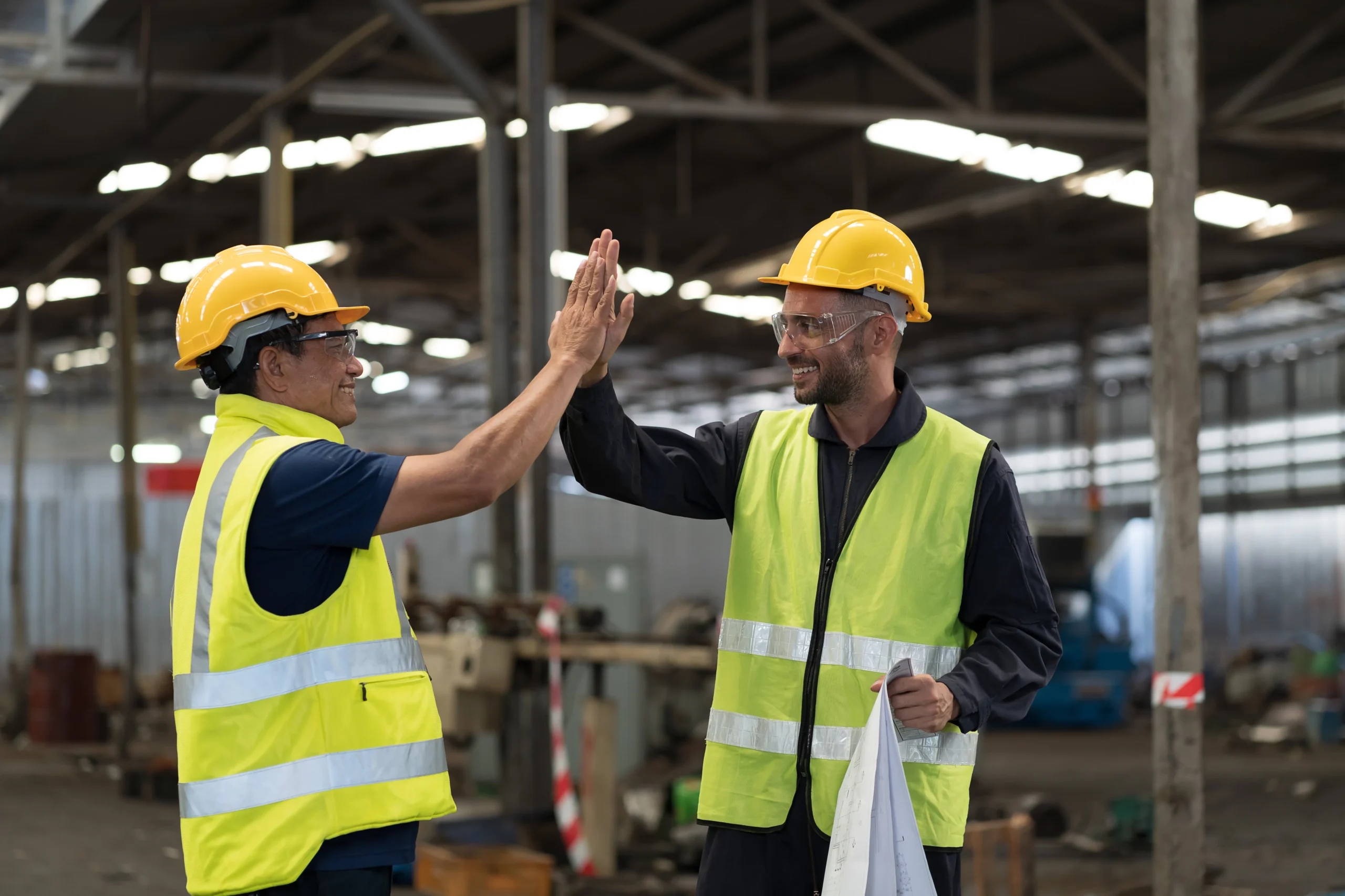 Two industrial workers in safety vests and hard hats giving a high five on the job site after completing a task safely. 