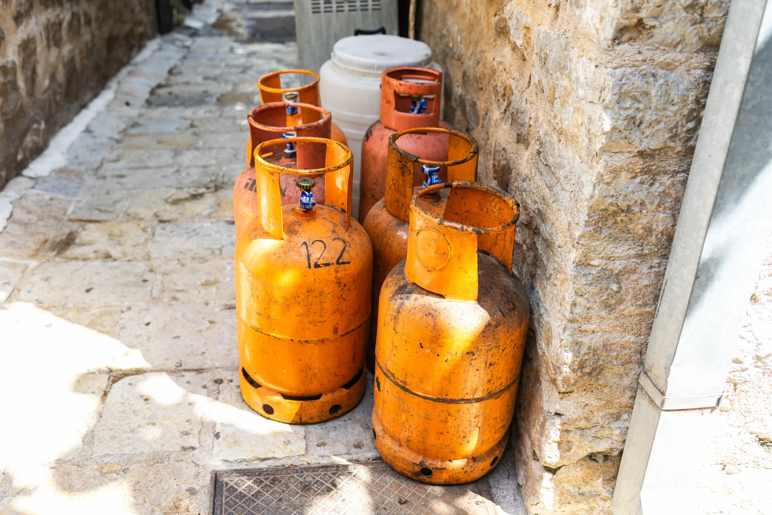 Multiple orange propane gas cylinders are stored outdoors against a stone wall, showing signs of rust and improper handling, posing a potential workplace safety hazard.