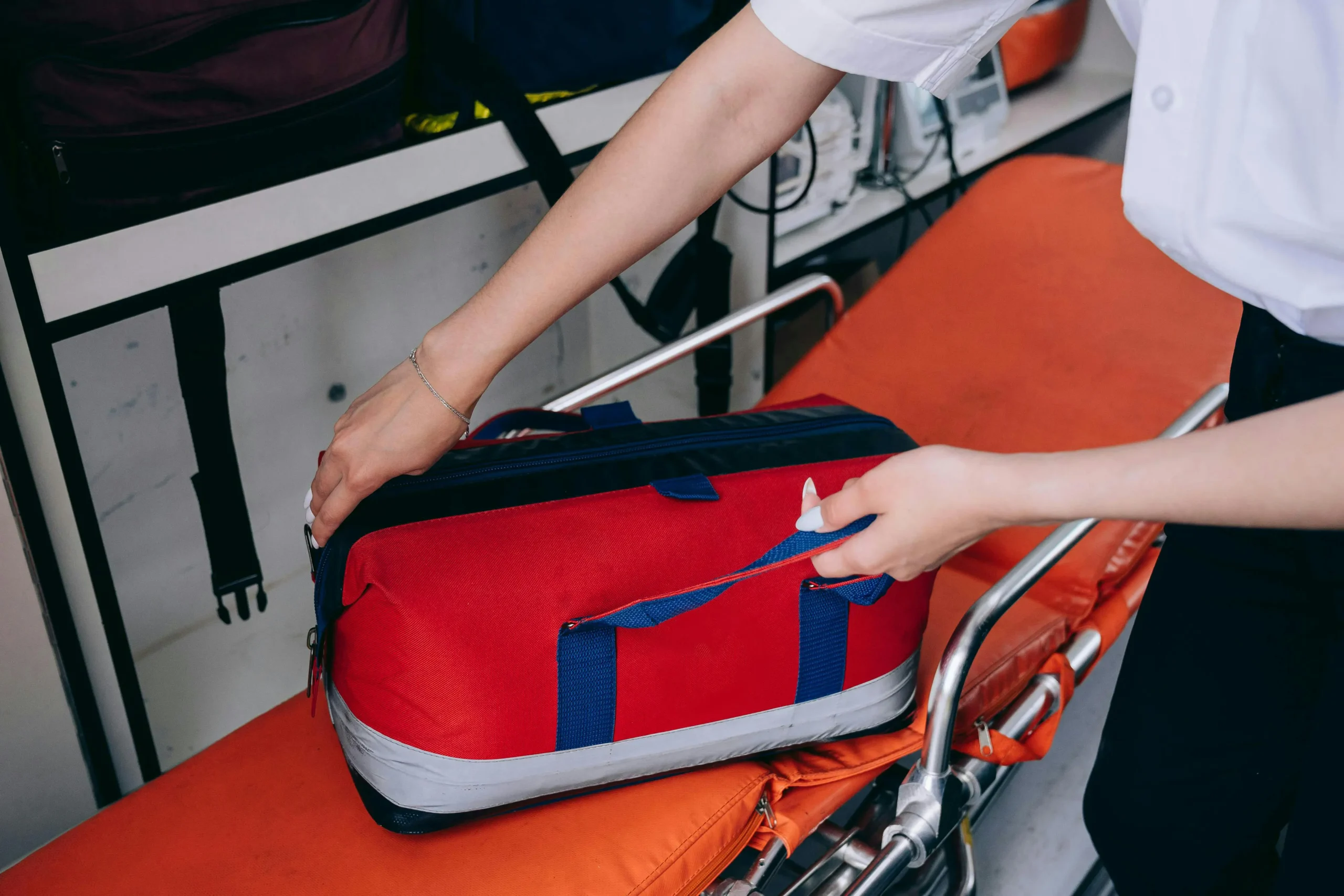First aid attendant opening a red first aid kit beside a stretcher in a medical setting.