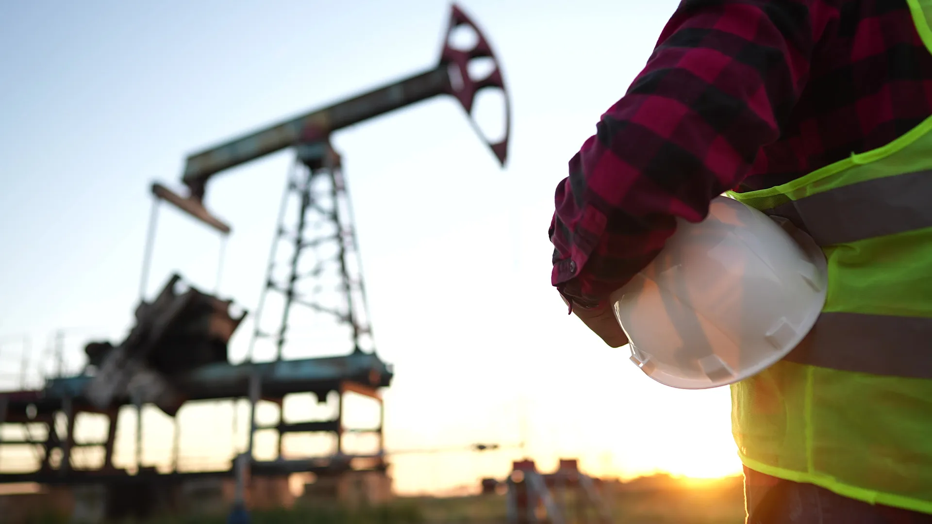 An oilfield worker in a high-visibility vest holding a hard hat at sunset with an oil pump jack in the background.
