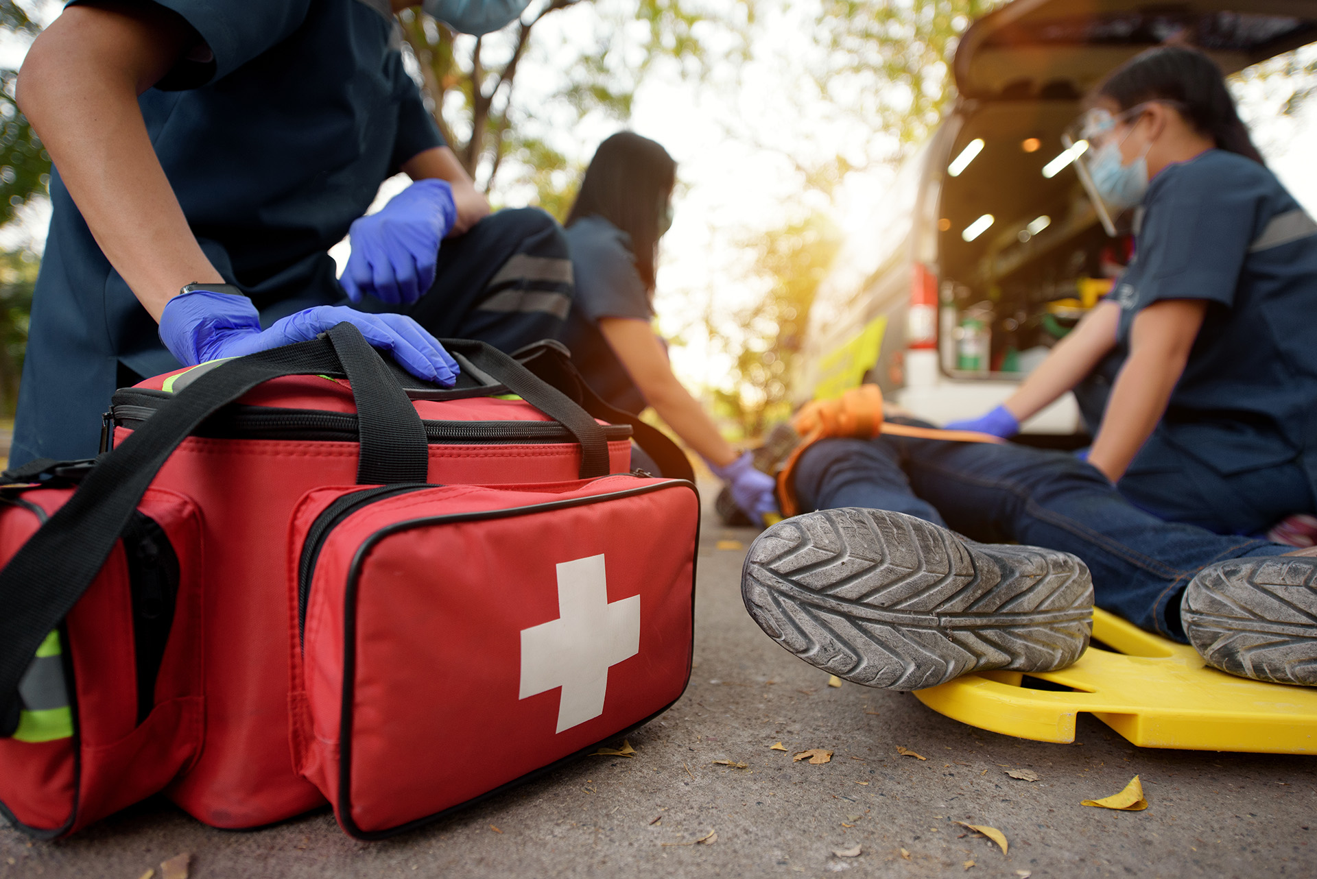Paramedics providing first aid to an injured person on a stretcher beside an ambulance with a medical kit in the foreground.