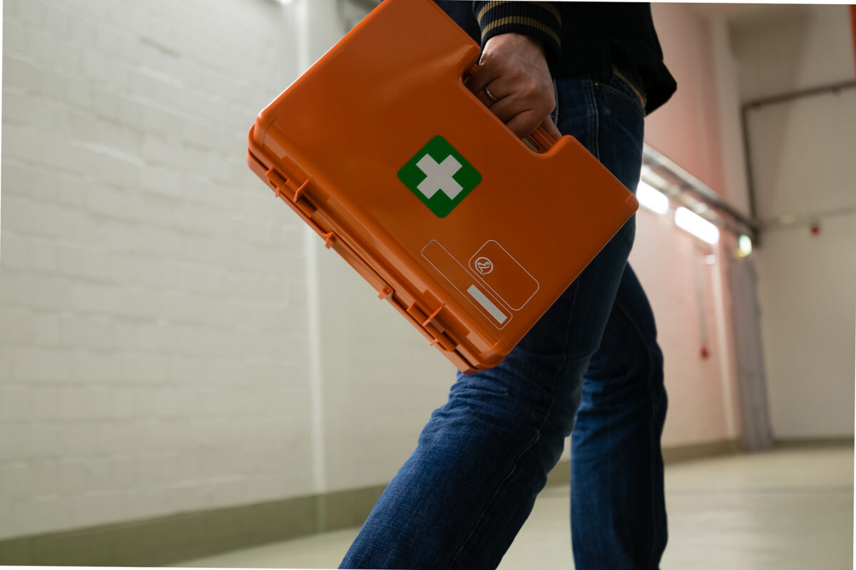 Person carrying an orange first aid kit with a white cross symbol while walking down a hallway.