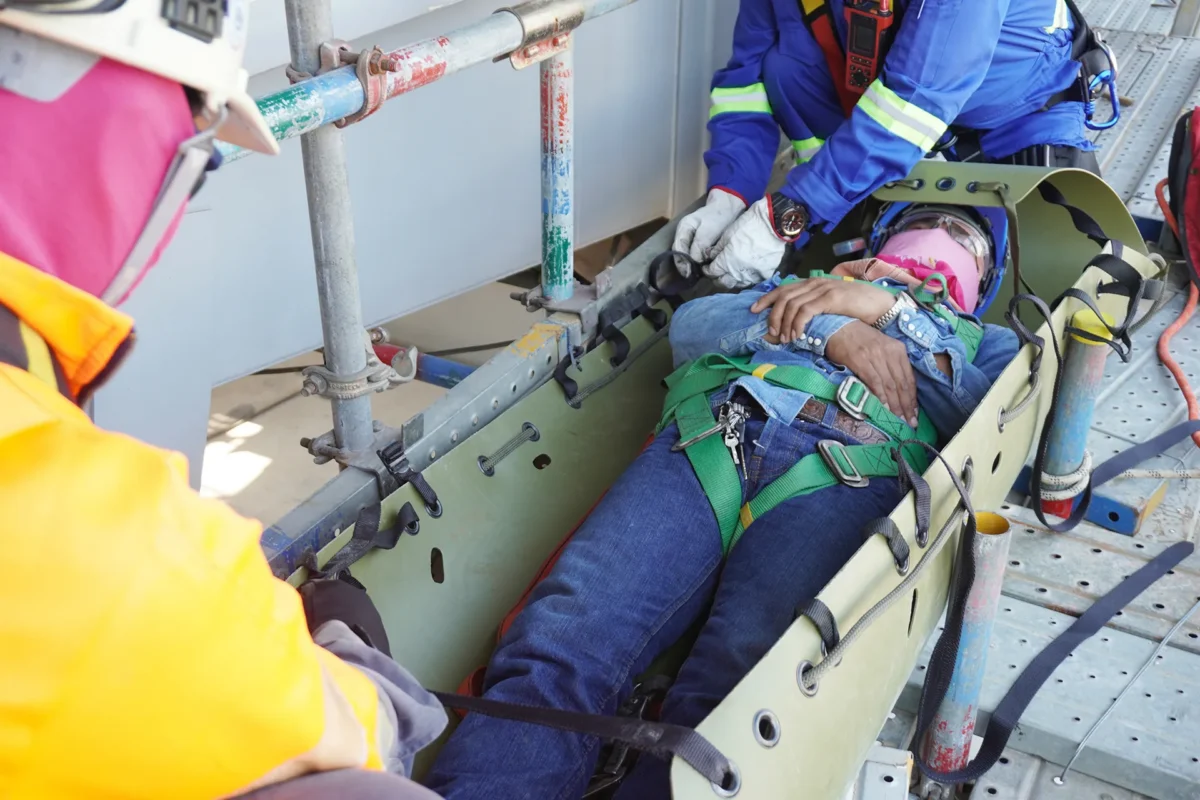 Industrial workers in high-visibility gear are packaging an injured worker onto a stretcher on an oilfield worksite