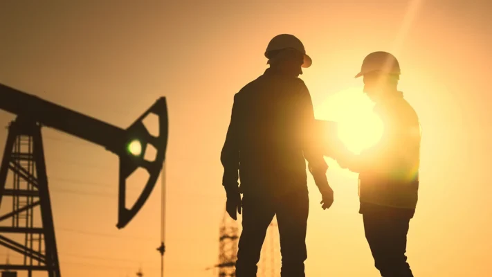 Two oilfield workers in hard hats stand in silhouette against a sunset sky beside a pump jack on an oil and gas worksite.