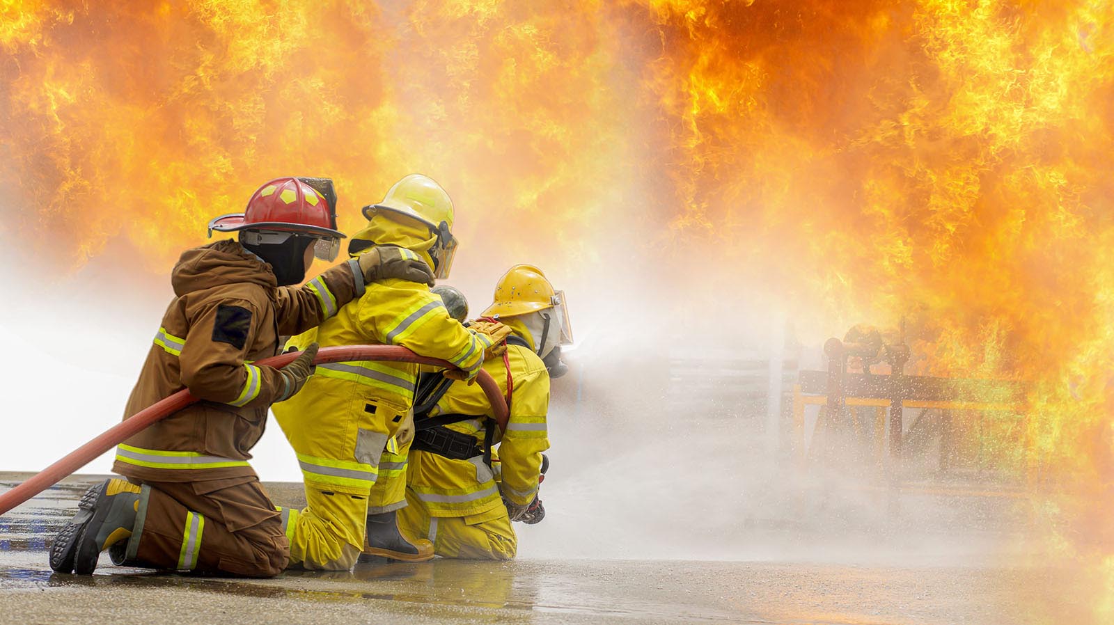 Firefighters in full protective gear kneel together while directing a high-pressure water hose at a large industrial fire, with towering flames and heavy spray filling the scene.