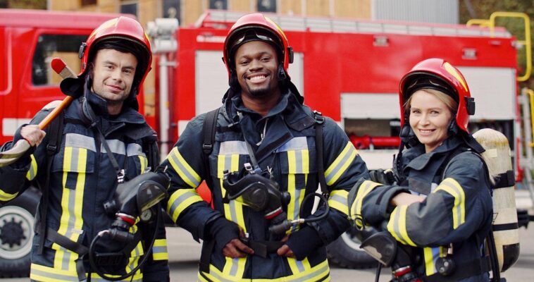 Three firefighters in full protective gear and helmets stand smiling in front of a fire truck, holding breathing apparatus masks.