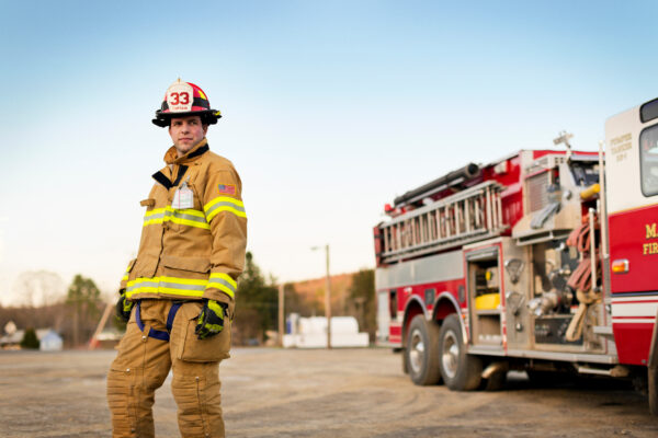 Firefighter in yellow turnout gear and helmet marked 33 stands beside a red fire truck, conveying readiness and calm on a clear day.