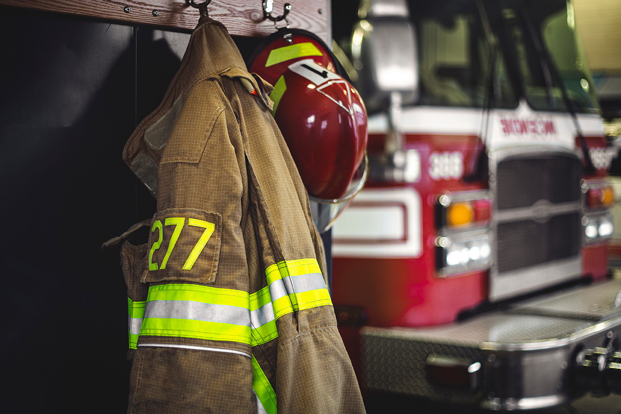 Firefighter turnout gear and helmet hanging inside a fire station, with a fire truck in the background.